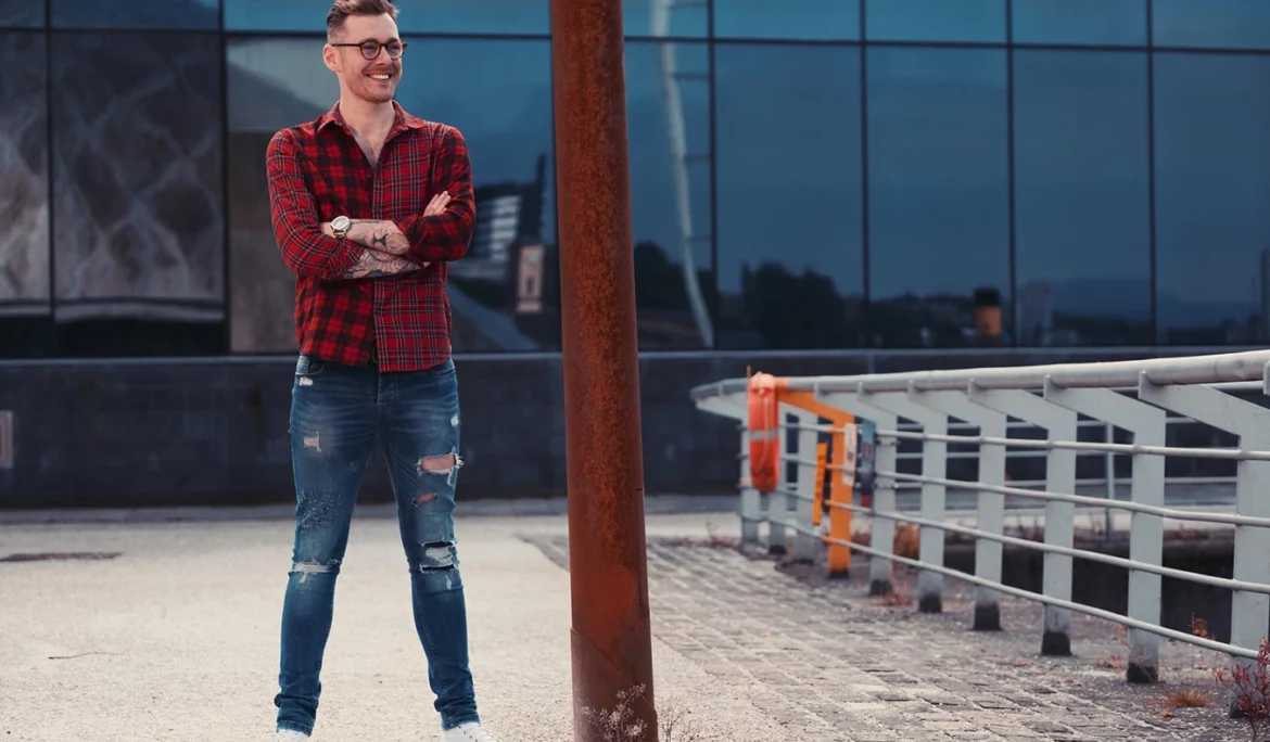 Male photographed in a red checked shirt and ripped blue jeans, standing looking happy and smiling with crossed arms. Photographed at Pacific Quay on a sunny summer afternoon. In the background are reflections of the Glasgow Science centre in the building windows and to the right the white metal railings by the waterside.