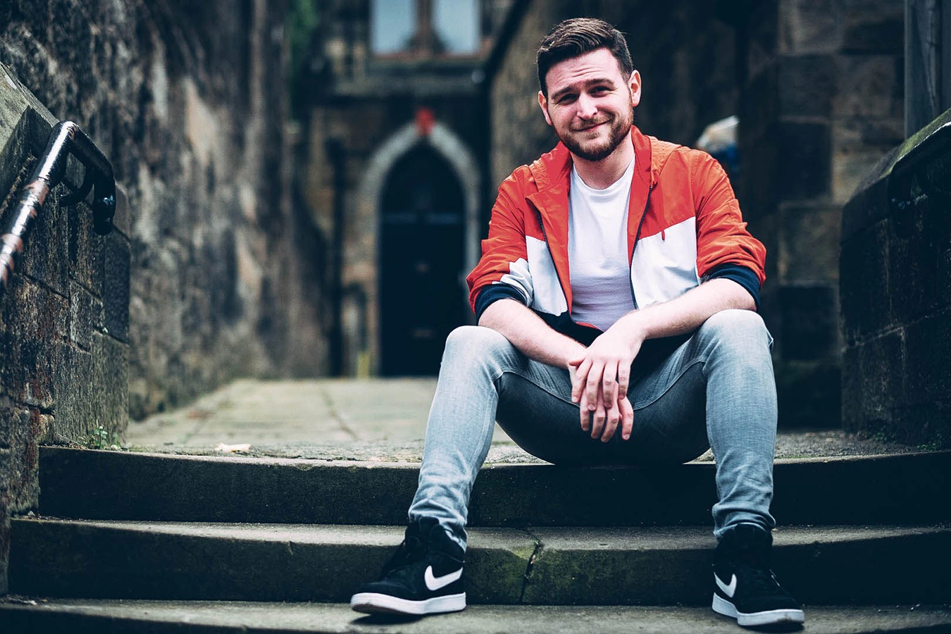 A Scottish actor, dancer and director, Andi Denny sits on stone steps outside a victorian building in the West End of Glasgow. He smiles at the camera. He is wearing a white and red light jacket and blue jeans.