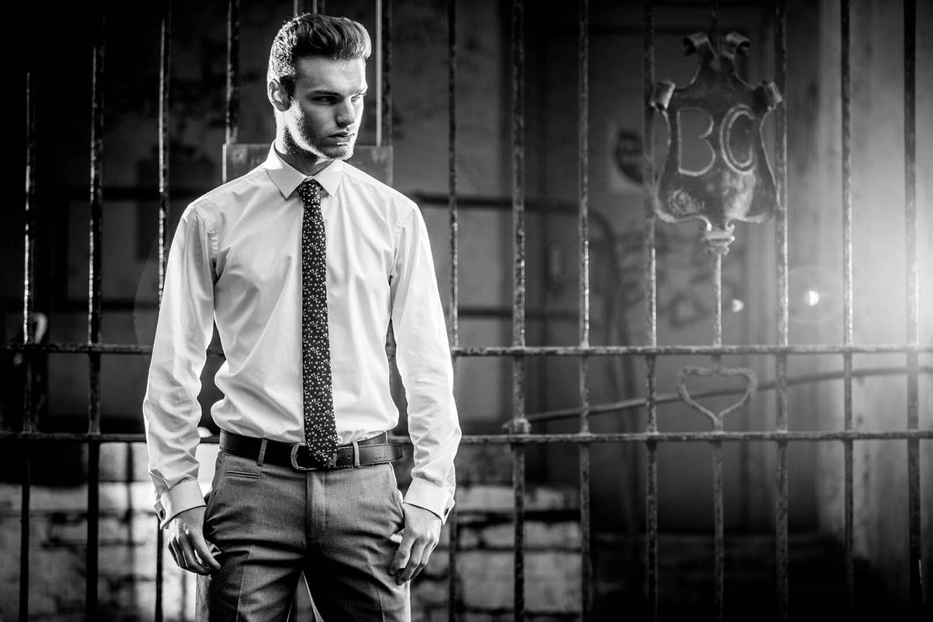 A black and white shot of a smartly dressed young male, wearing white shirt and patterned tie looks to camera right. He is standing in front of a wrought iron gate, and is lit by bright light source out of frame on the right hand side, Personal Branding Shoots