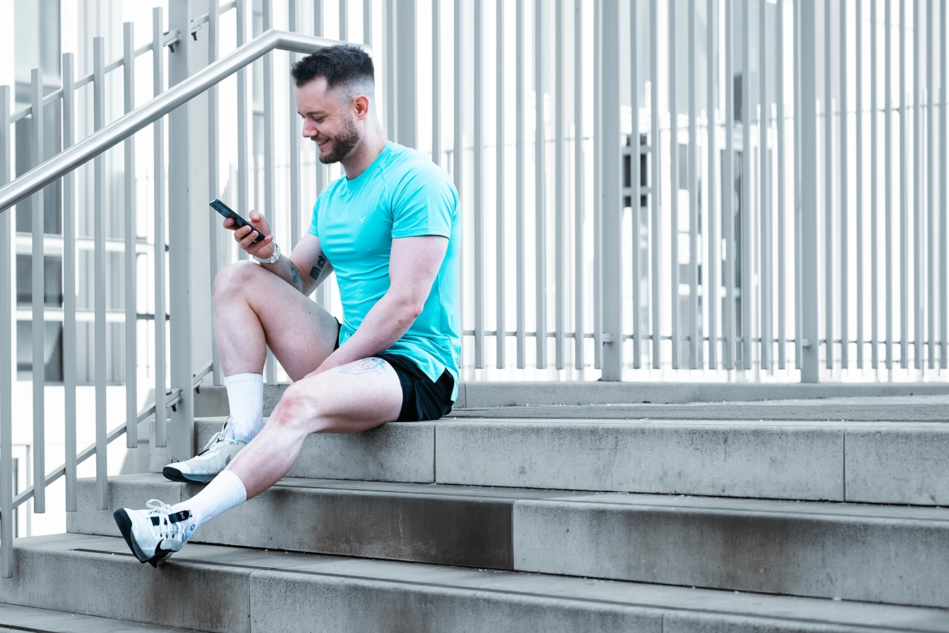 A young male sits on stone steps outside Glasgow City College in the Merchant City. He is smiling while scrolling through his phone. He wears a turquoise t-shirt and black shorts, Branding Shoots