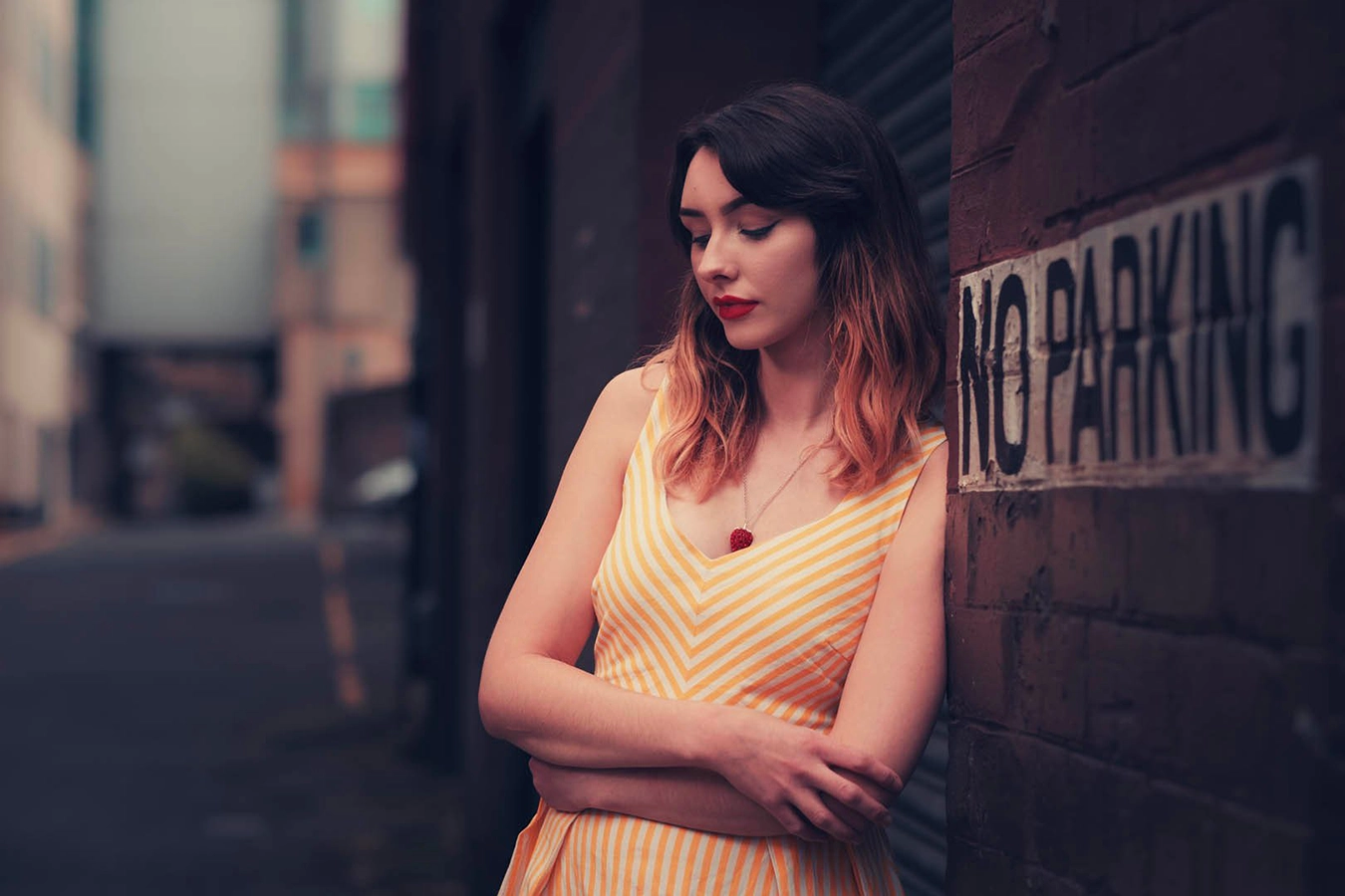 A young lady in a yellow dress poses leaning against a metal garage shutter door. She is standing in an alley, eyes closed looking reflective. To her left hand side a No Parking sign is toward the camera.