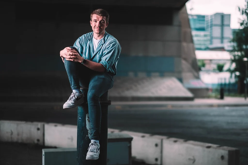 Actor Steven, is photographed perched atop a bollard, in a relaxed informal pose under Glasgow's Kingston Bridge. The city skyline can be seen in the background.