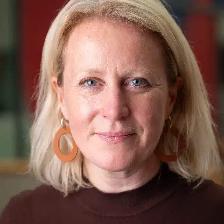 Headshot of a female business executive, looking directly to camera. She has blonde hair with a dark plum woolen top. She has circular orange earrings and looks confident and assured.